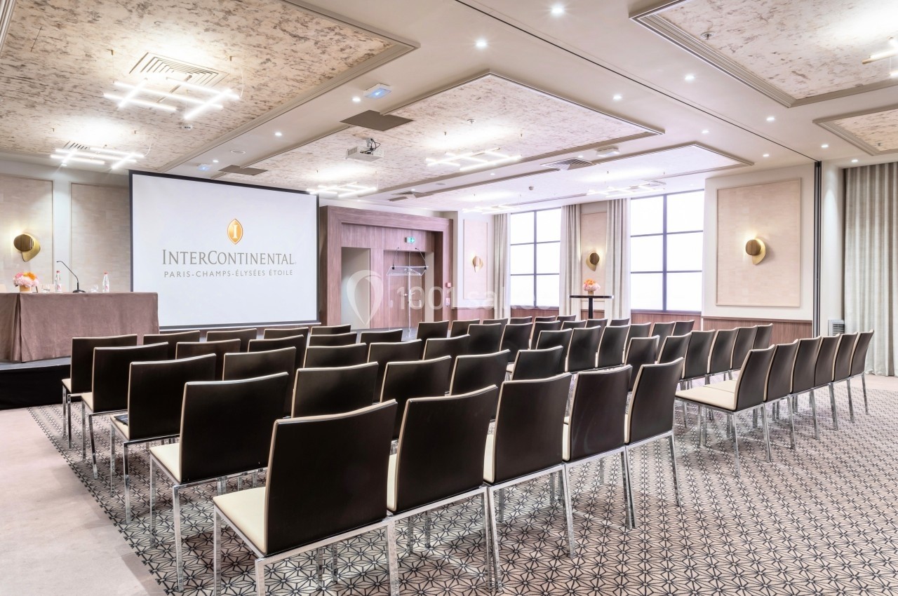 Salle de conférence lumineuse avec rangées de chaises, écran de projection et table pour présentateurs.