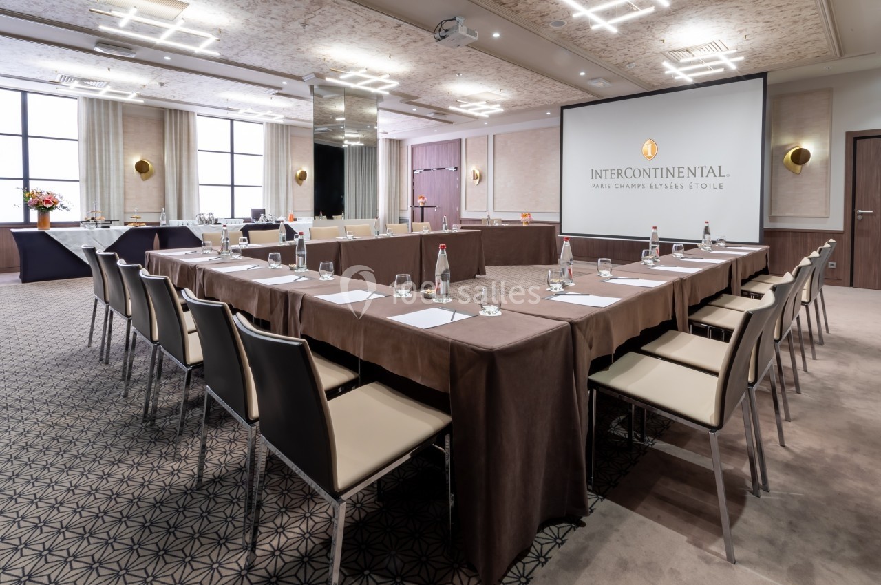Salle de réunion lumineuse avec tables disposées en U, chaises beige, bouteilles d'eau et écran de projection.