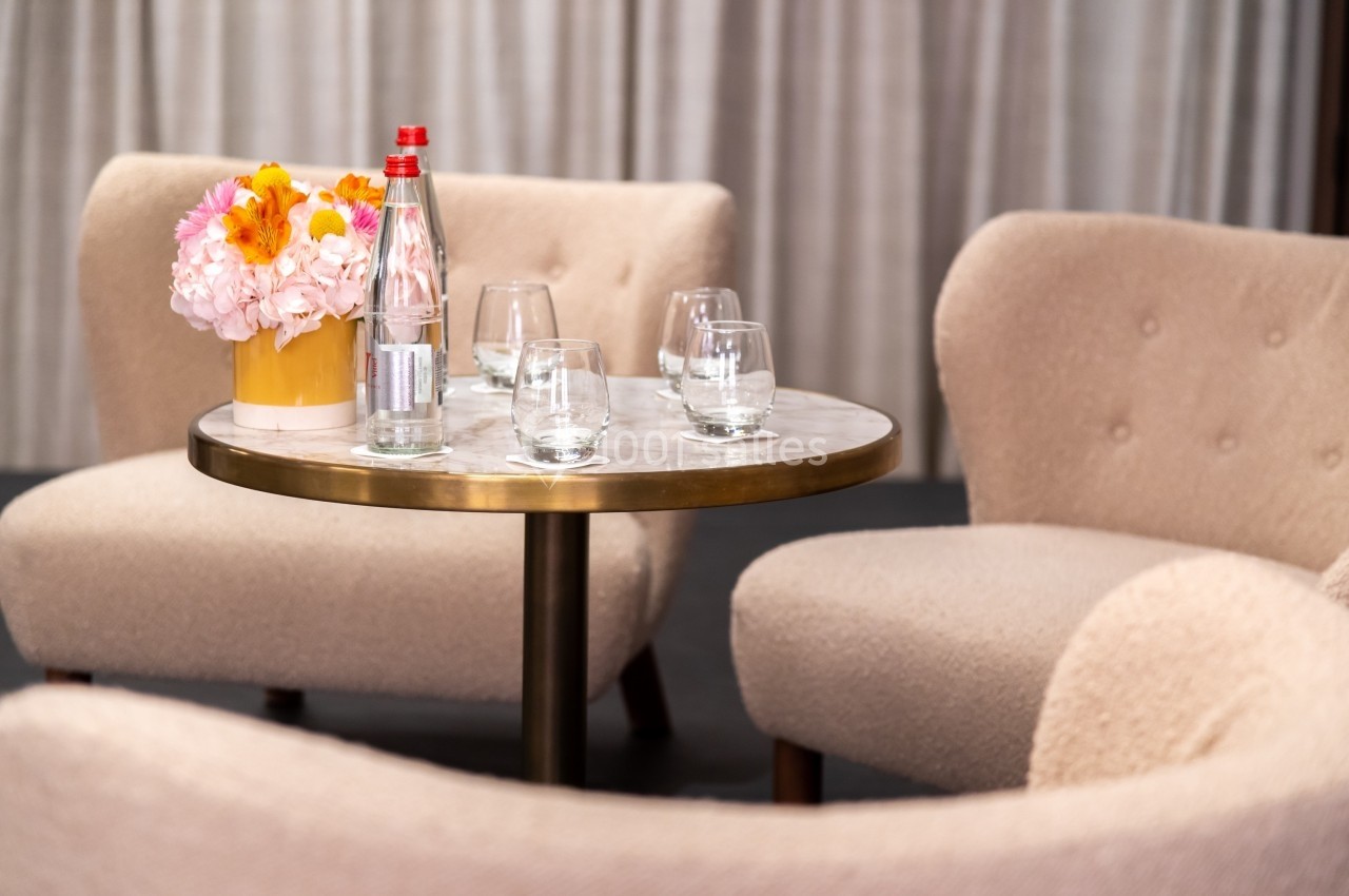 Table ronde en marbre entourée de fauteuils beiges, avec un vase de fleurs, une bouteille d'eau et des verres.