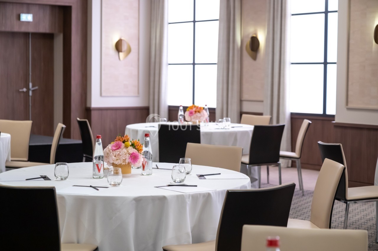 Salle de réception avec tables rondes dressées, chaises beige et noires, et décorations florales au centre.