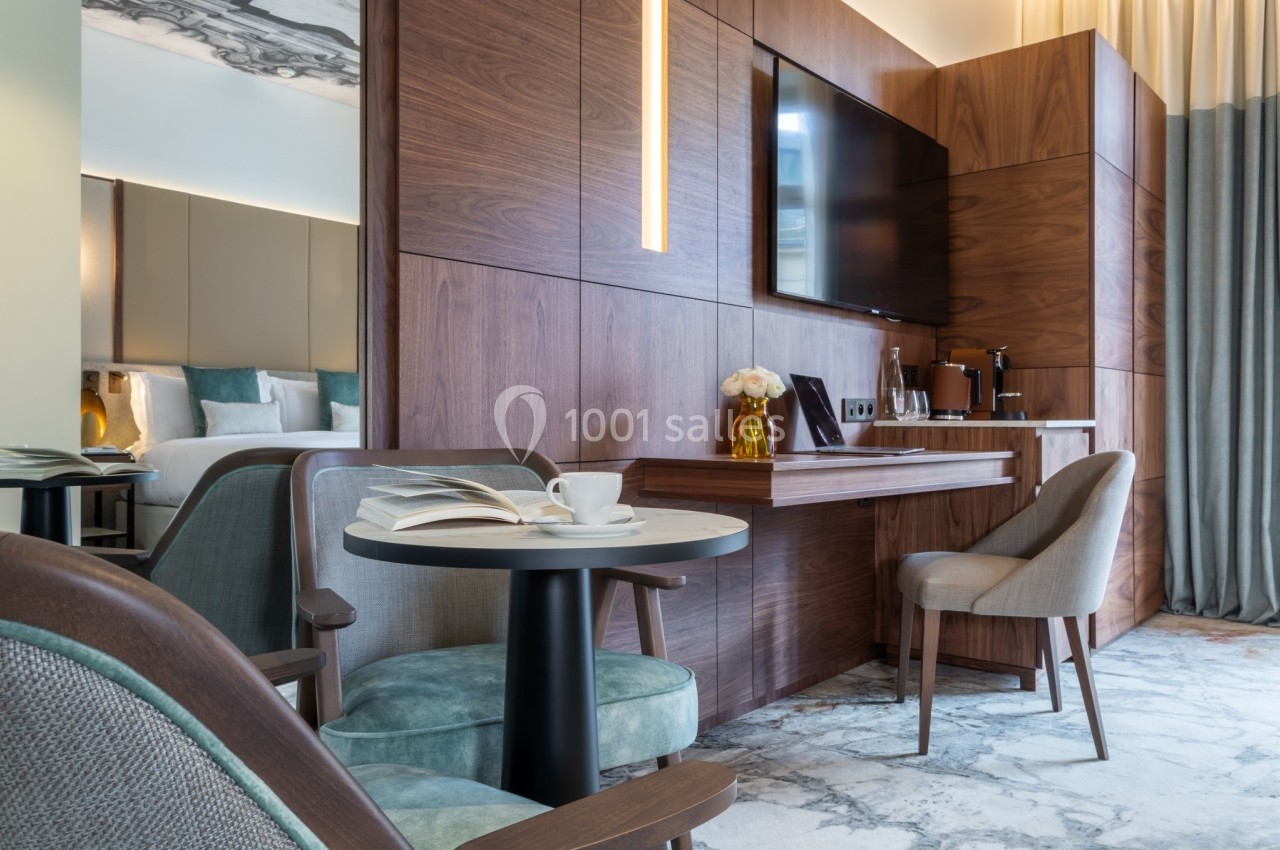 Coin salon d'une chambre d'hôtel avec table, chaises, télévision murale, lit en arrière-plan et éclairage chaleureux.