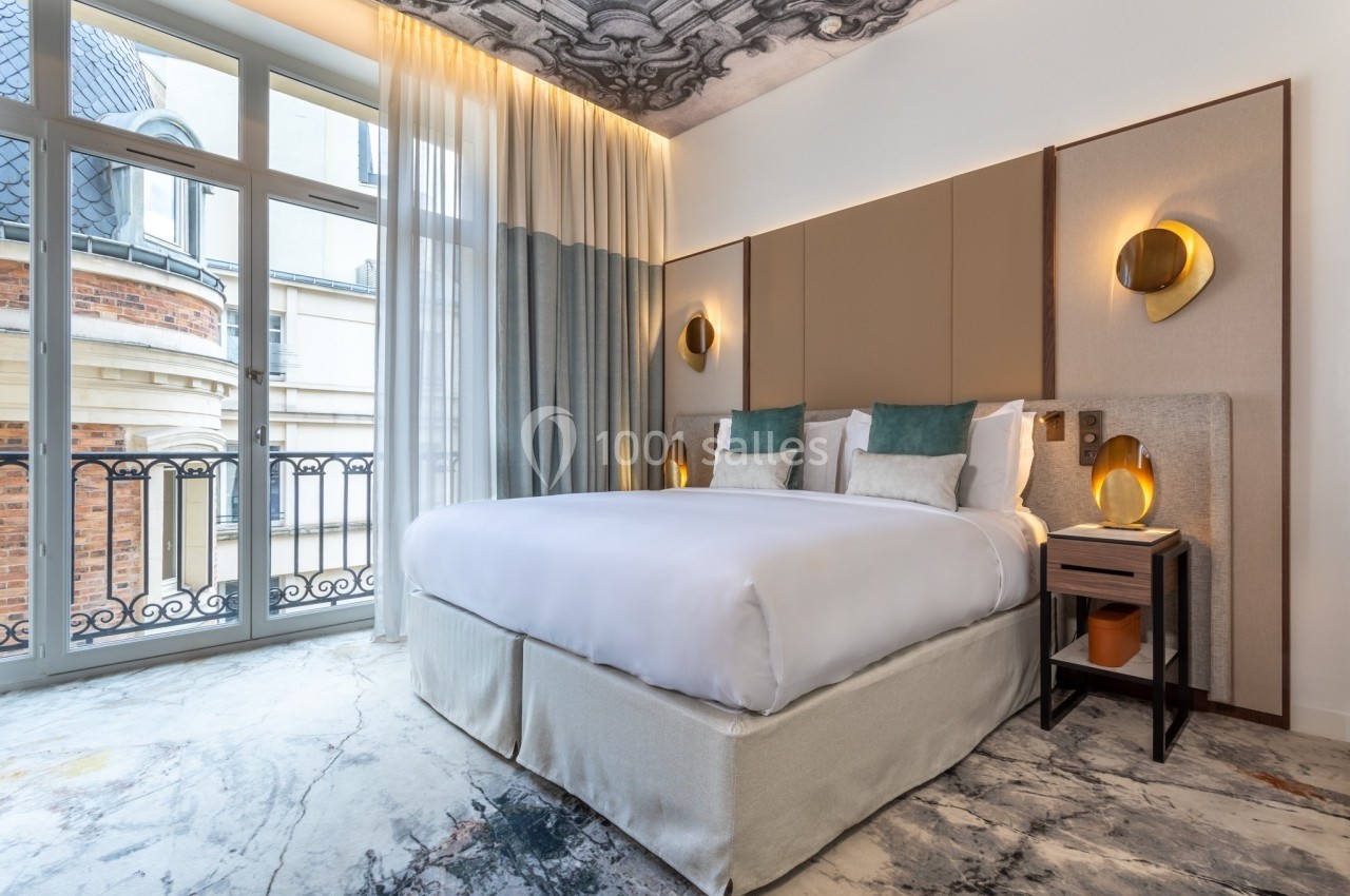 Chambre d'hôtel lumineuse avec un grand lit, des lampes murales dorées, un plafond décoré et une vue sur des bâtiments…