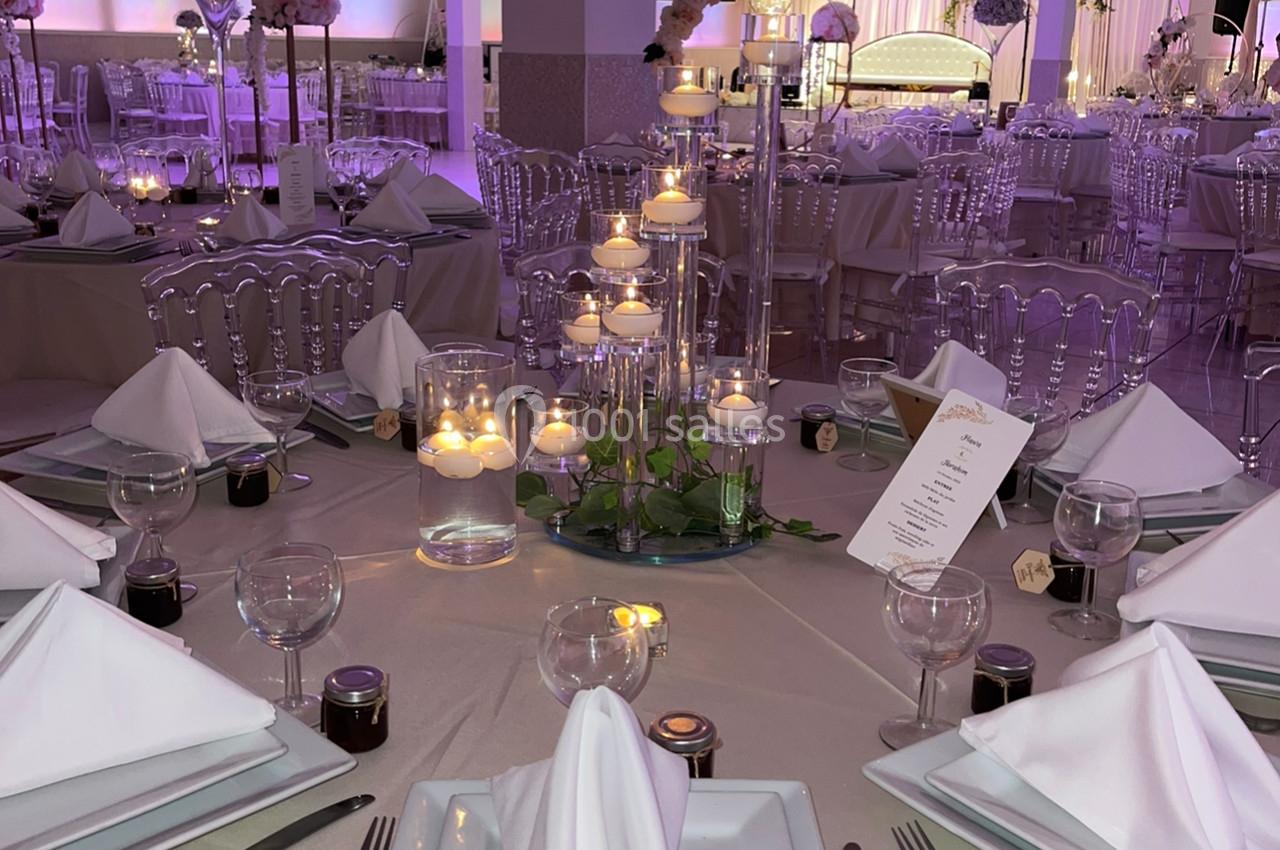 Salle de réception décorée avec des tables élégantes, nappes beiges, bougies et éclairage violet tamisé.