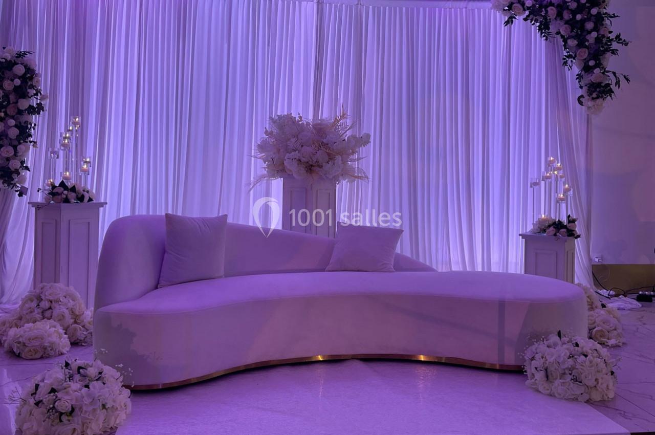 Canapé courbé violet clair devant des rideaux plissés, entouré de fleurs blanches et de chandeliers décoratifs.