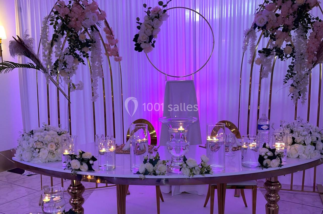 Table décorée pour un événement avec fleurs blanches, bougies et arche dorée sur fond de rideaux blancs éclairés en violet.