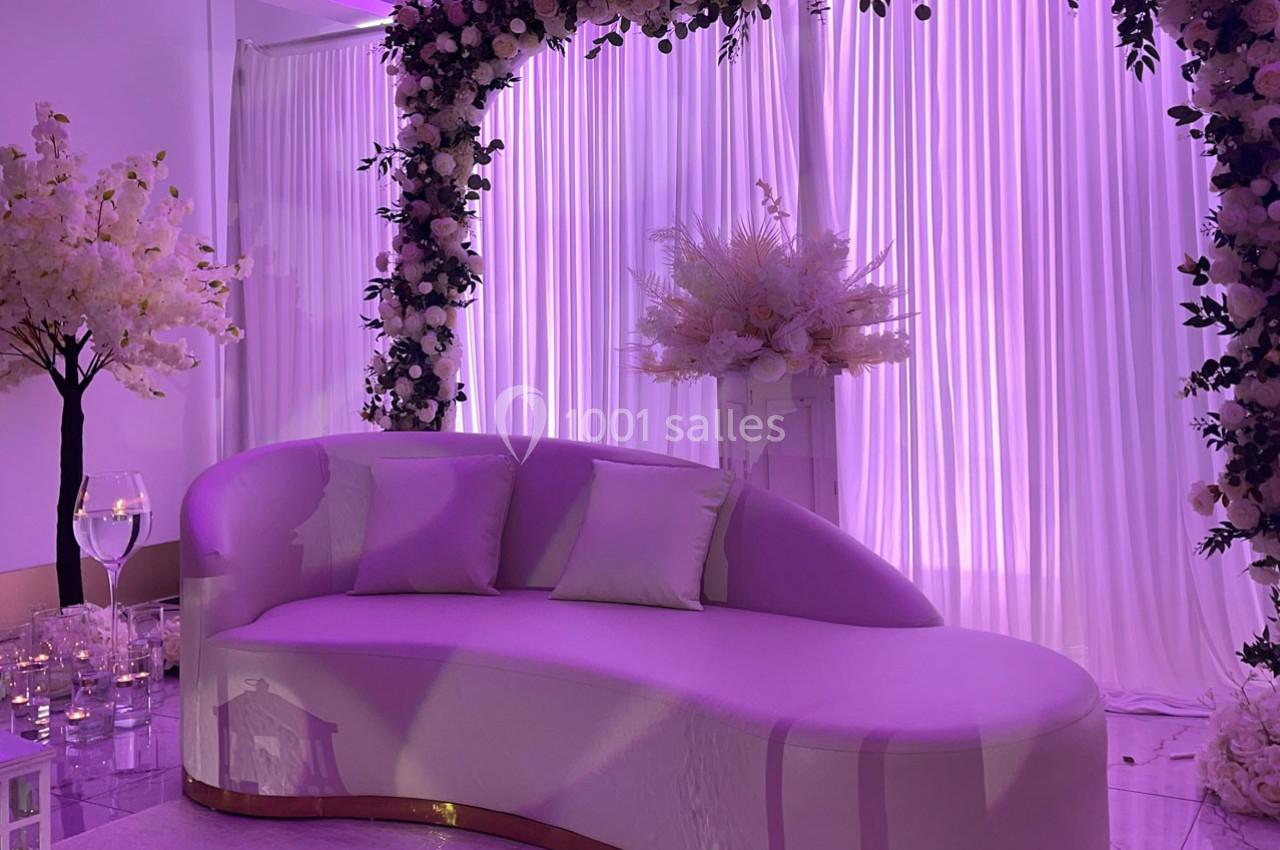 Canapé courbé blanc devant des rideaux lumineux violets, décoré d'un arc floral et d'arrangements floraux élégants.