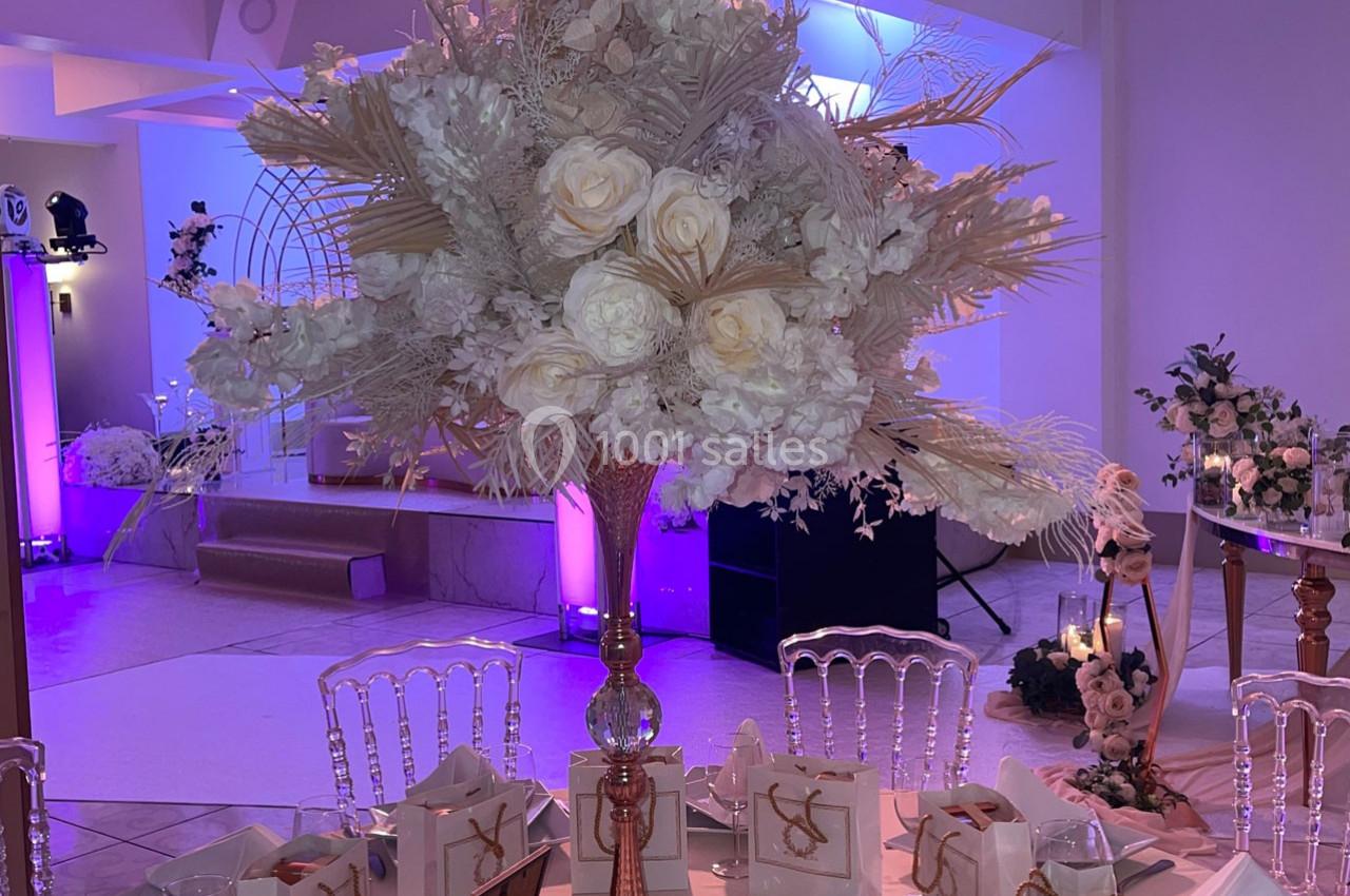 Centre de table floral avec roses blanches et feuillage beige, dans une salle décorée de lumières violettes.