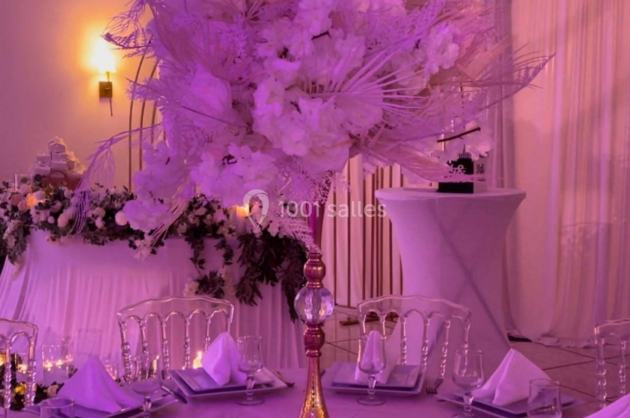 Centre de table floral avec des fleurs blanches et roses sur une table dressée dans une salle de réception éclairée.