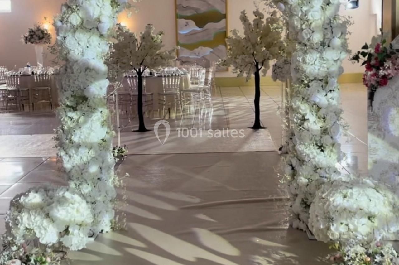 Arche de mariage décorée de fleurs blanches, entourée de chaises transparentes et d'arbres artificiels.
