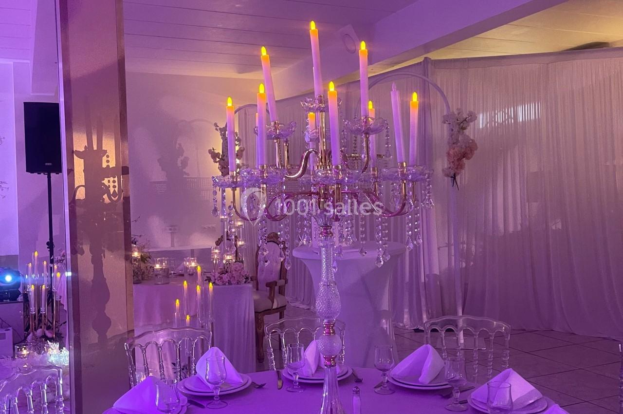 Centre de table avec chandelier doré et bougies LED, entouré de tables dressées dans une salle éclairée en violet.