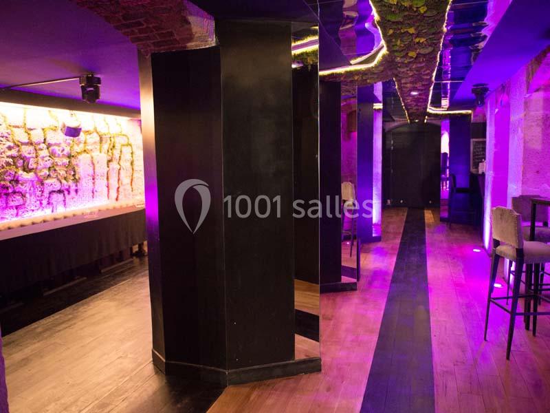 Salle intérieure avec murs en pierre, éclairage violet, piliers noirs et sol en bois.