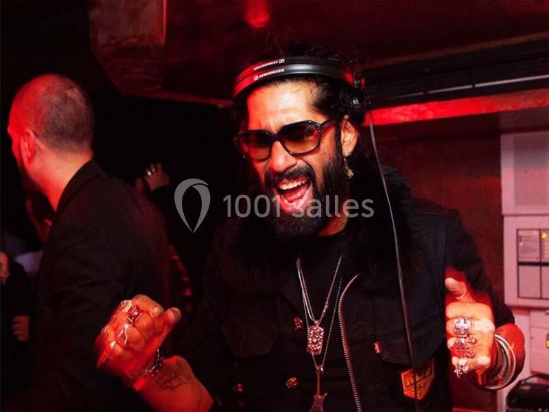 Un DJ barbu portant des lunettes de soleil mixe de la musique dans une ambiance lumineuse rouge.