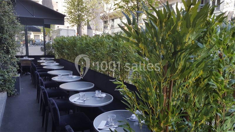 Terrasse de restaurant avec tables rondes dressées, entourée de haies végétales dans un environnement urbain.