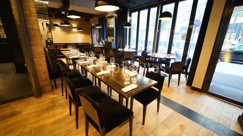 Salle de restaurant lumineuse avec tables en bois dressées, chaises noires et grandes baies vitrées donnant sur l'extérieur.