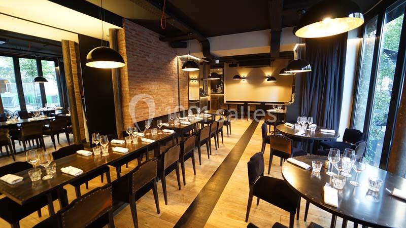 Salle de restaurant moderne avec tables en bois, chaises noires, grandes baies vitrées et éclairage suspendu.
