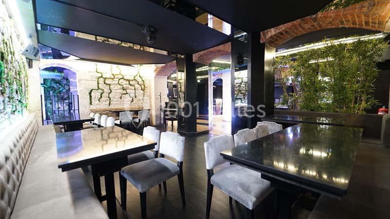 Salle de restaurant moderne avec tables en bois sombre, chaises rembourrées et murs décorés de plantes et briques apparentes.