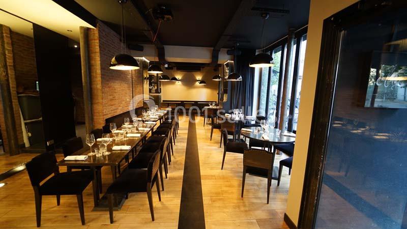 Salle de restaurant moderne avec tables dressées, chaises noires, éclairage suspendu et parquet en bois clair.