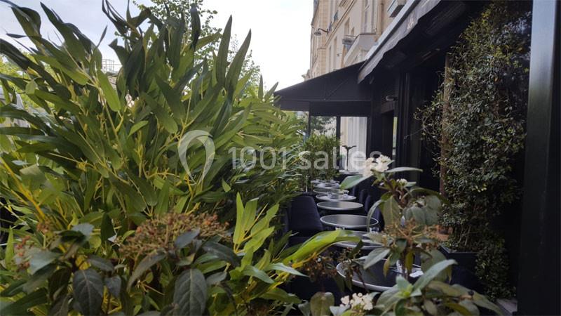 Terrasse extérieure bordée de plantes vertes, avec des tables et des chaises alignées le long d'un bâtiment.
