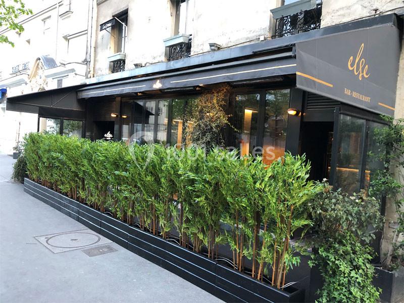 Façade d'un restaurant avec terrasse entourée de plantes vertes, situé le long d'un trottoir en ville.
