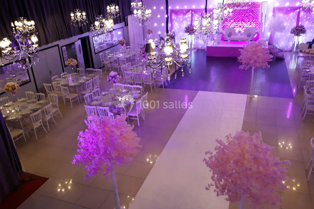 Salle de réception décorée avec des tables, des chaises blanches, des lustres et des arbres roses sous un éclairage violet.