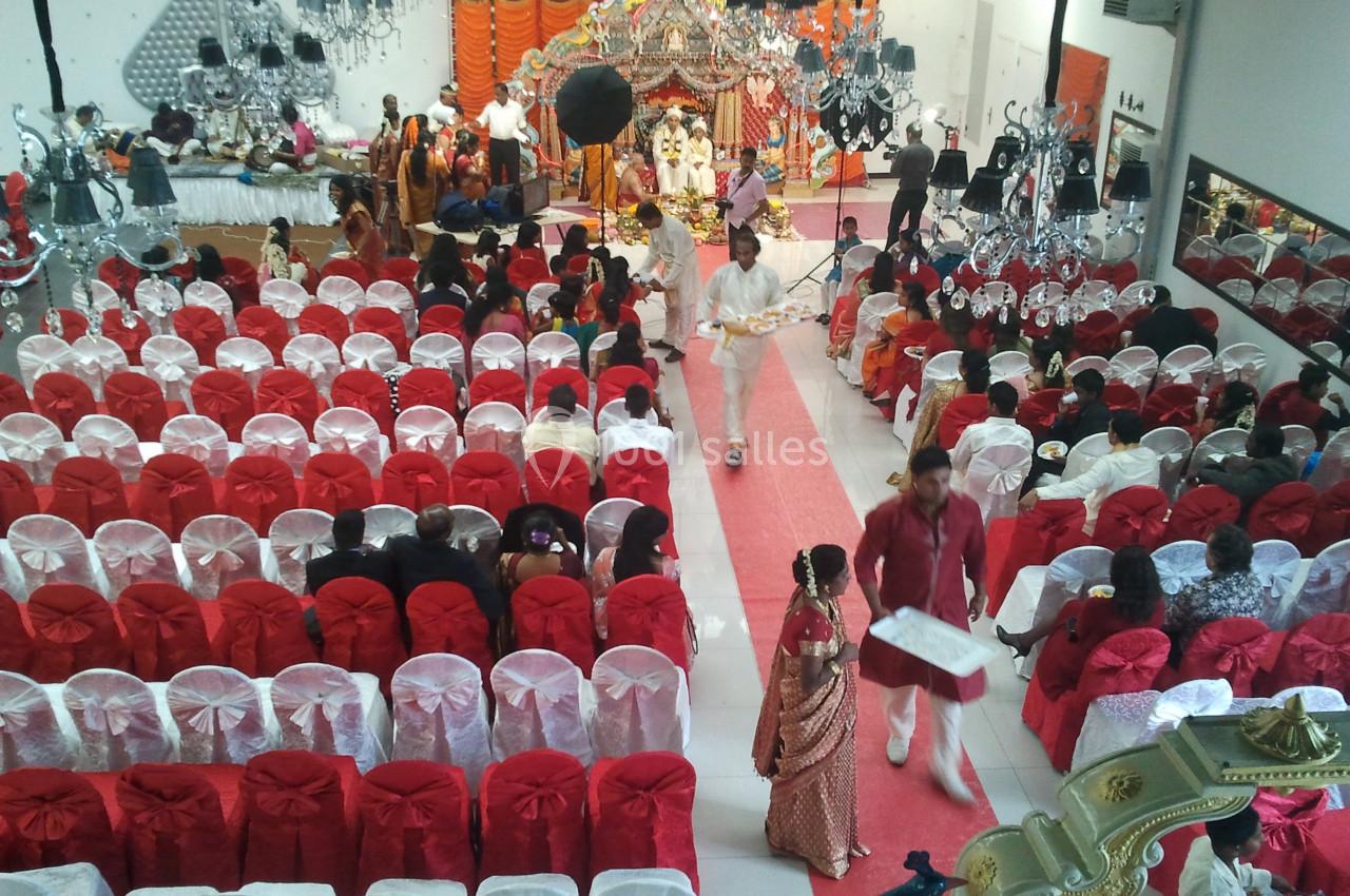 Salle décorée pour un mariage avec invités assis, allée centrale rouge et mariés sous une arche ornée.
