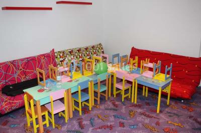 Salle colorée avec petites tables et chaises pour enfants, entourées de canapés aux motifs vifs sur un tapis décoré.