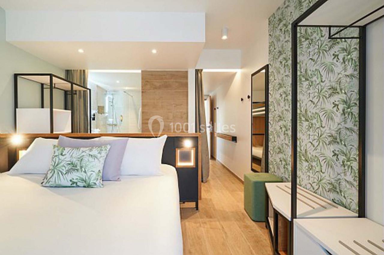 Chambre moderne avec lit double, décor végétal, éclairage doux et vue sur une salle de bain ouverte en arrière-plan.