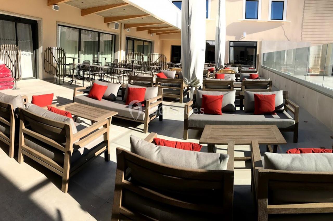 Terrasse ensoleillée avec mobilier en bois, coussins rouges et gris, tables basses et baies vitrées en arrière-plan.