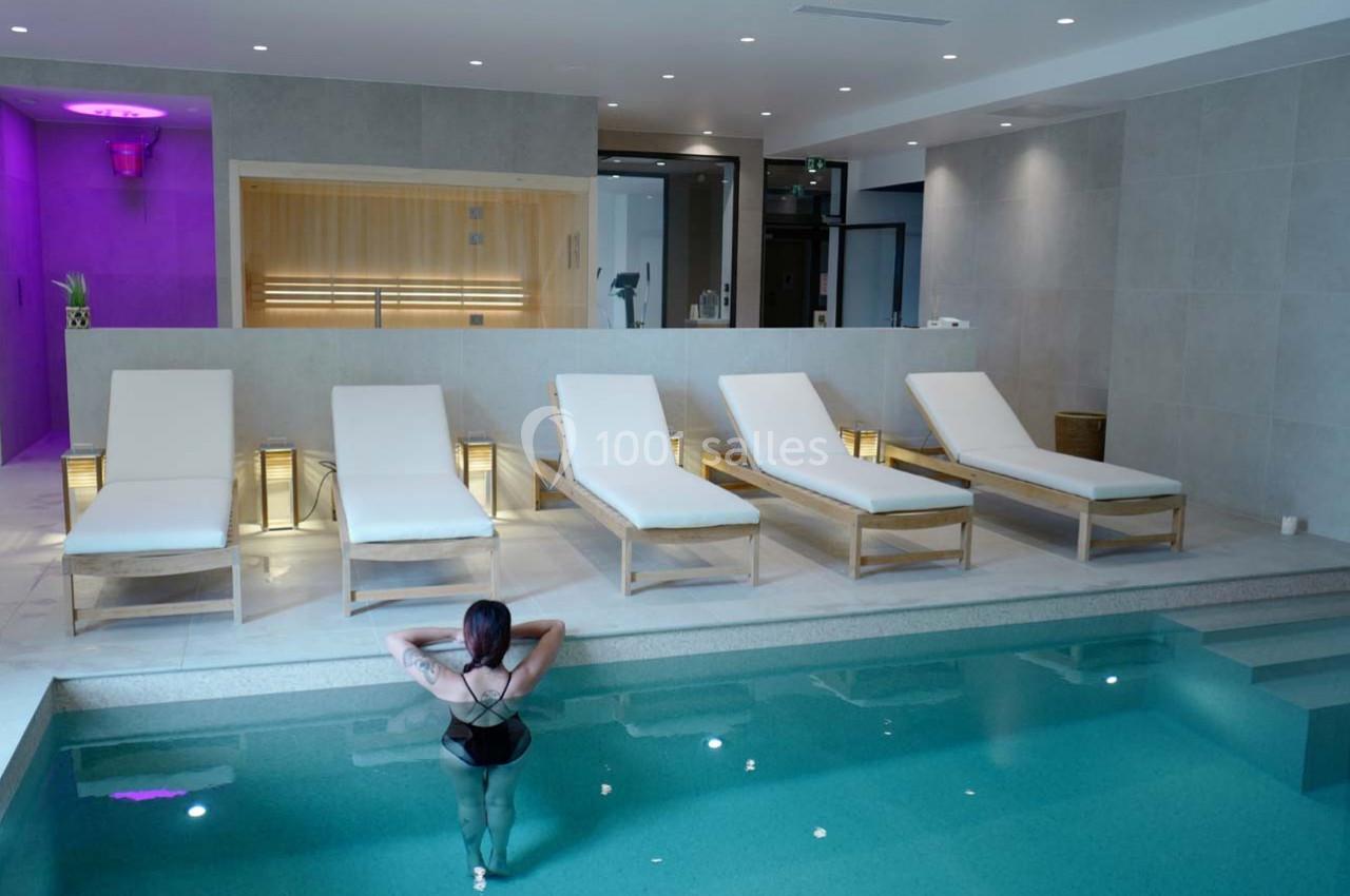 Femme debout dans une piscine intérieure, face à une rangée de chaises longues et une cabine sauna éclairée.