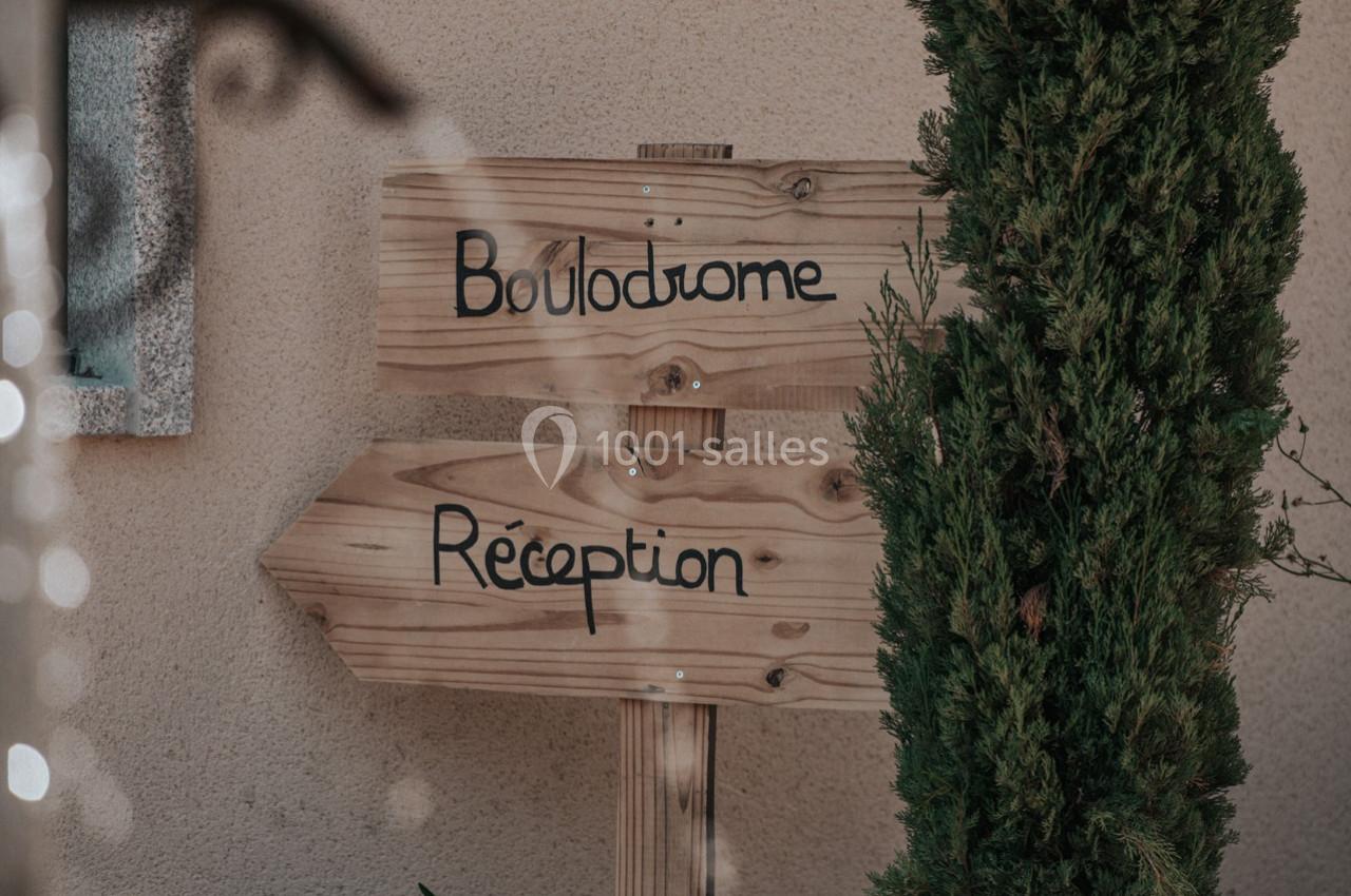 Panneaux en bois indiquant les directions pour le boulodrome et la réception, près d'un mur beige et d'un arbre.