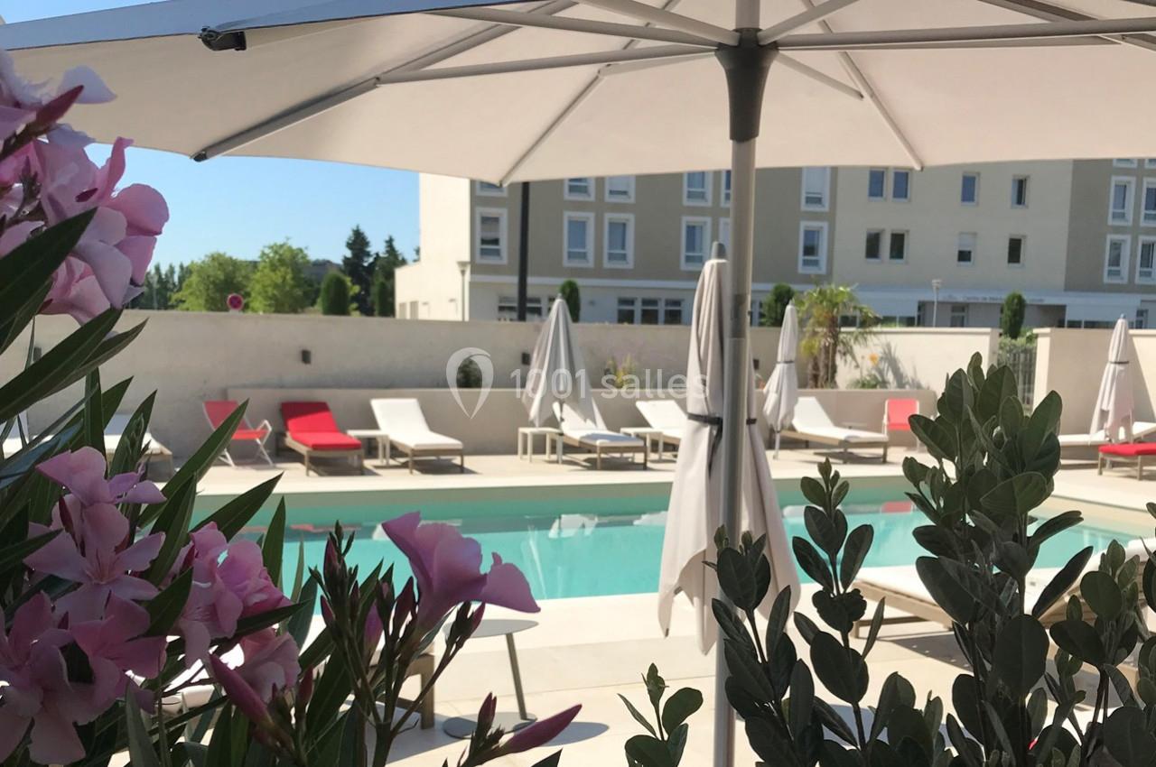 Piscine extérieure entourée de chaises longues et parasols, avec des fleurs au premier plan et un bâtiment en arrière-plan.