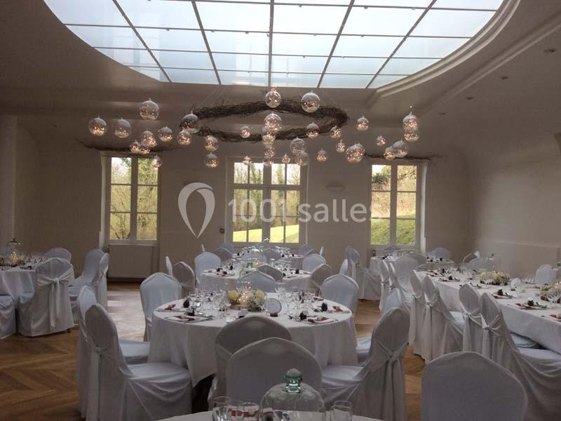 Salle de réception lumineuse avec tables rondes dressées, nappes blanches et décorations suspendues sous une verrière.