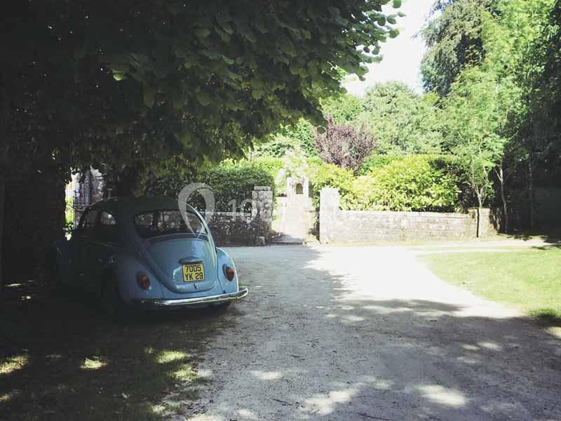 Location salle Audierne (Finistère) - Manoir de Menez Bras #18 Une voiture ancienne bleue garée à l'ombre d'un arbre près d'une allée bordée de verdure et d'un mur en pierre.