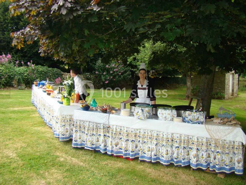 Location salle Audierne (Finistère) - Manoir de Menez Bras #21 Table dressée en extérieur sous des arbres, avec nappes décorées, plats variés et une personne en tenue traditionnelle.