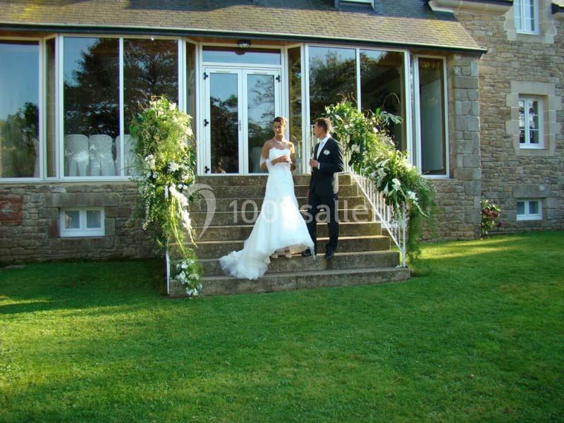 Location salle Audierne (Finistère) - Manoir de Menez Bras #19 Un couple en tenue de mariage pose sur les marches fleuries devant une maison en pierre avec de grandes fenêtres.