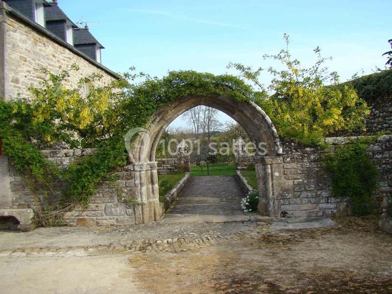 Location salle Audierne (Finistère) - Manoir de Menez Bras #17 Arche en pierre ancienne entourée de végétation, donnant sur un chemin bordé de murs et un paysage champêtre.