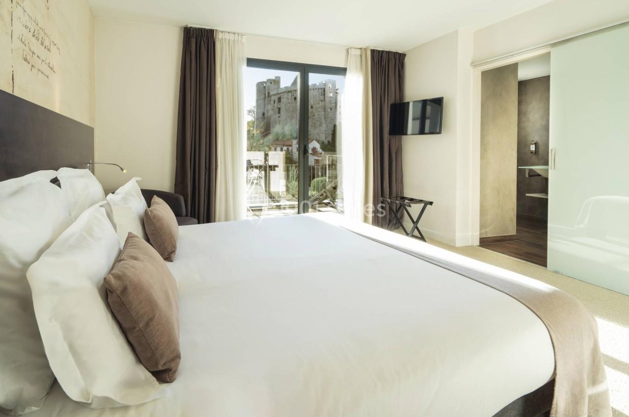 Chambre lumineuse avec lit double, grandes fenêtres donnant sur un château, et salle de bain attenante.