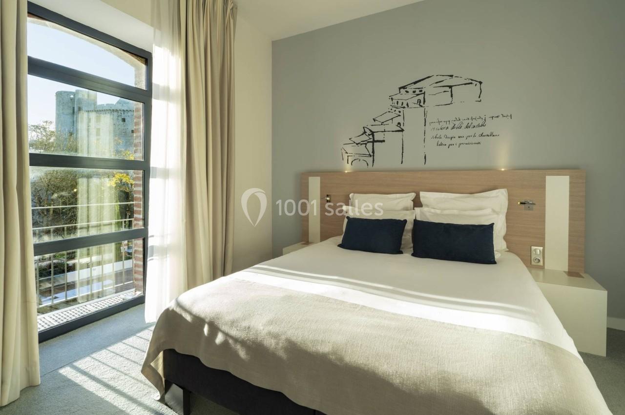 Chambre lumineuse avec un lit double, deux coussins bleus, une grande fenêtre et une décoration murale minimaliste.