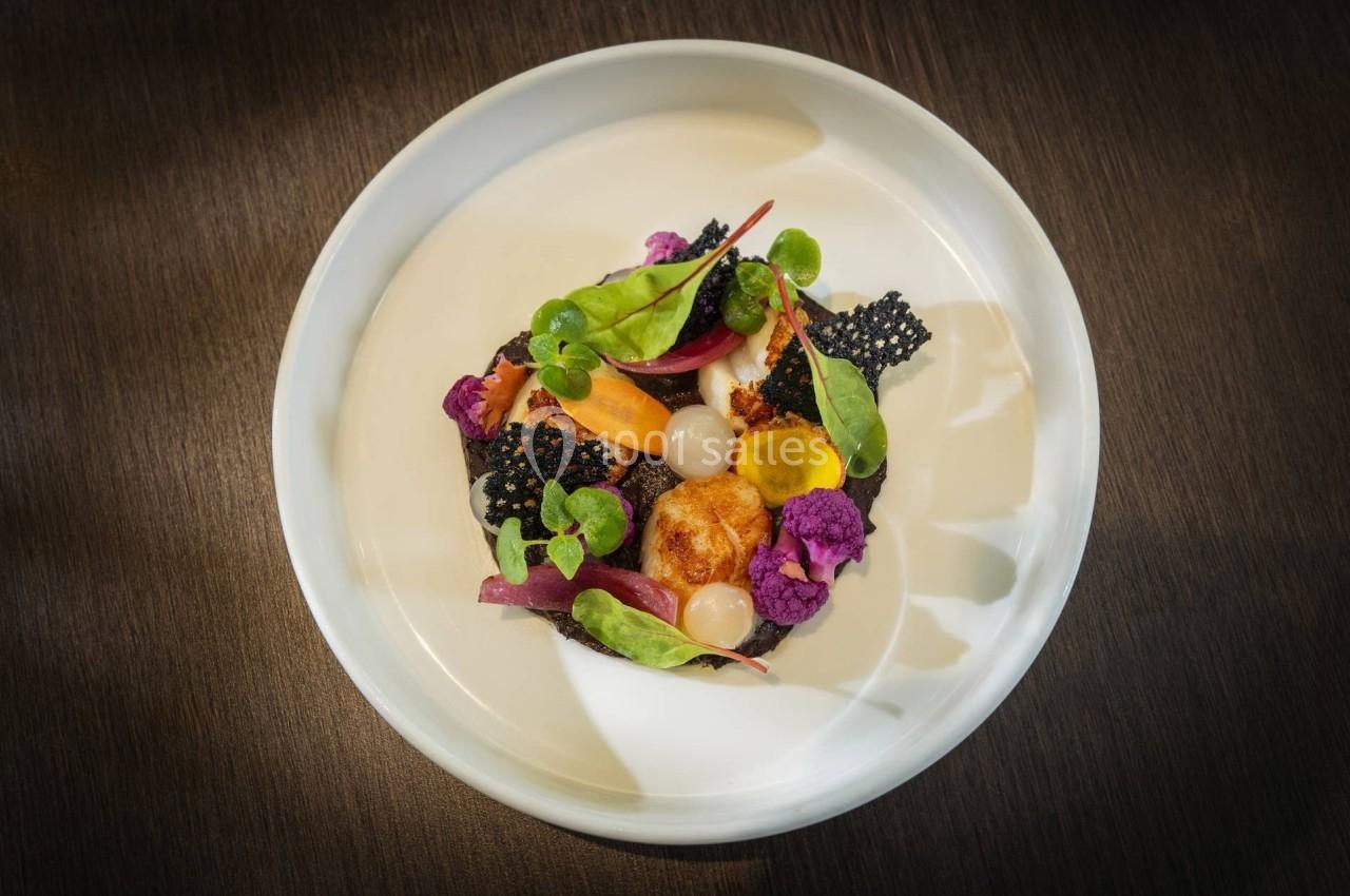 Assiette gastronomique avec légumes colorés, noix de Saint-Jacques et tuiles noires, présentée sur une table en bois sombre.