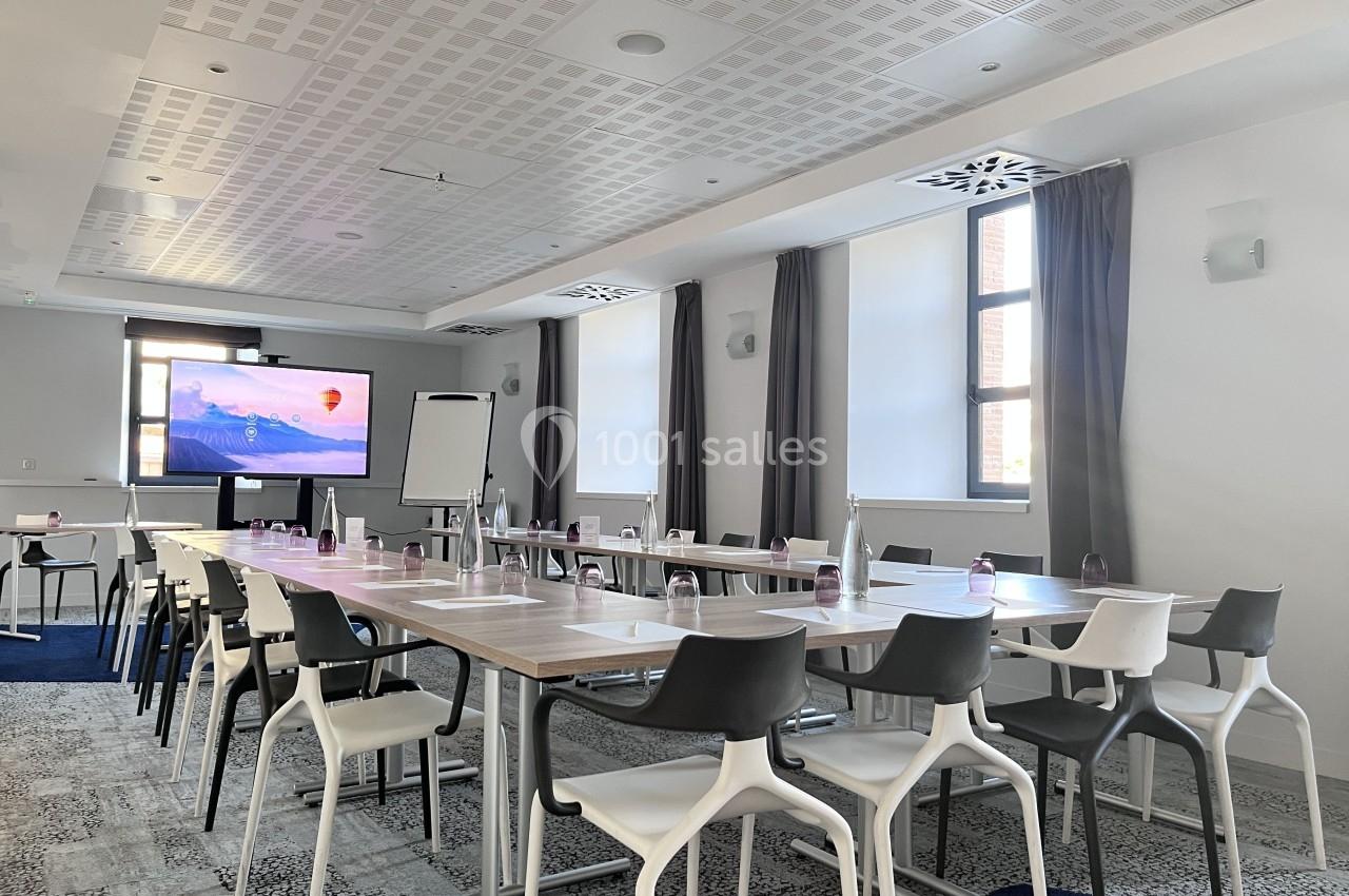 Salle de réunion lumineuse avec des tables disposées en U, chaises blanches et écran de présentation allumé.