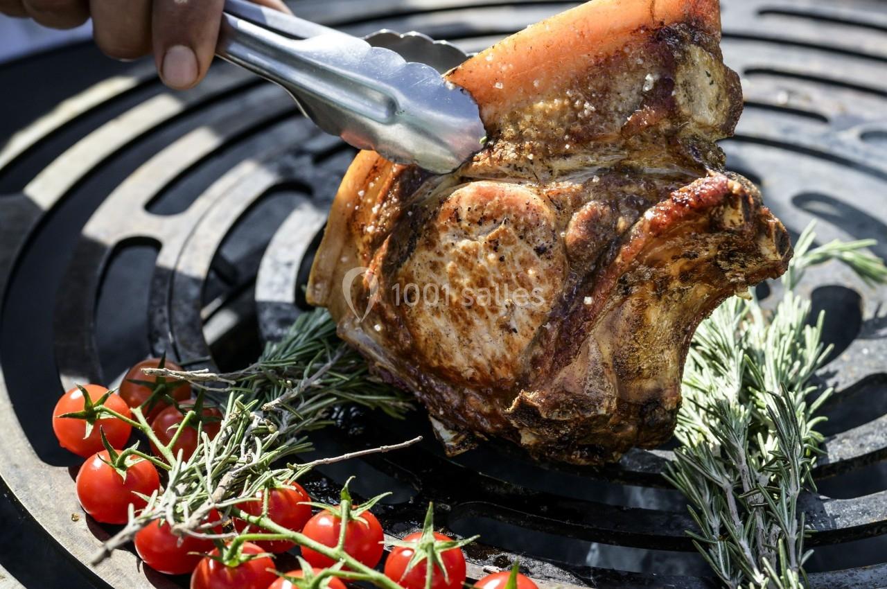 Viande grillée sur un barbecue avec des tomates cerises et des herbes fraîches, tenue par une pince en métal.
