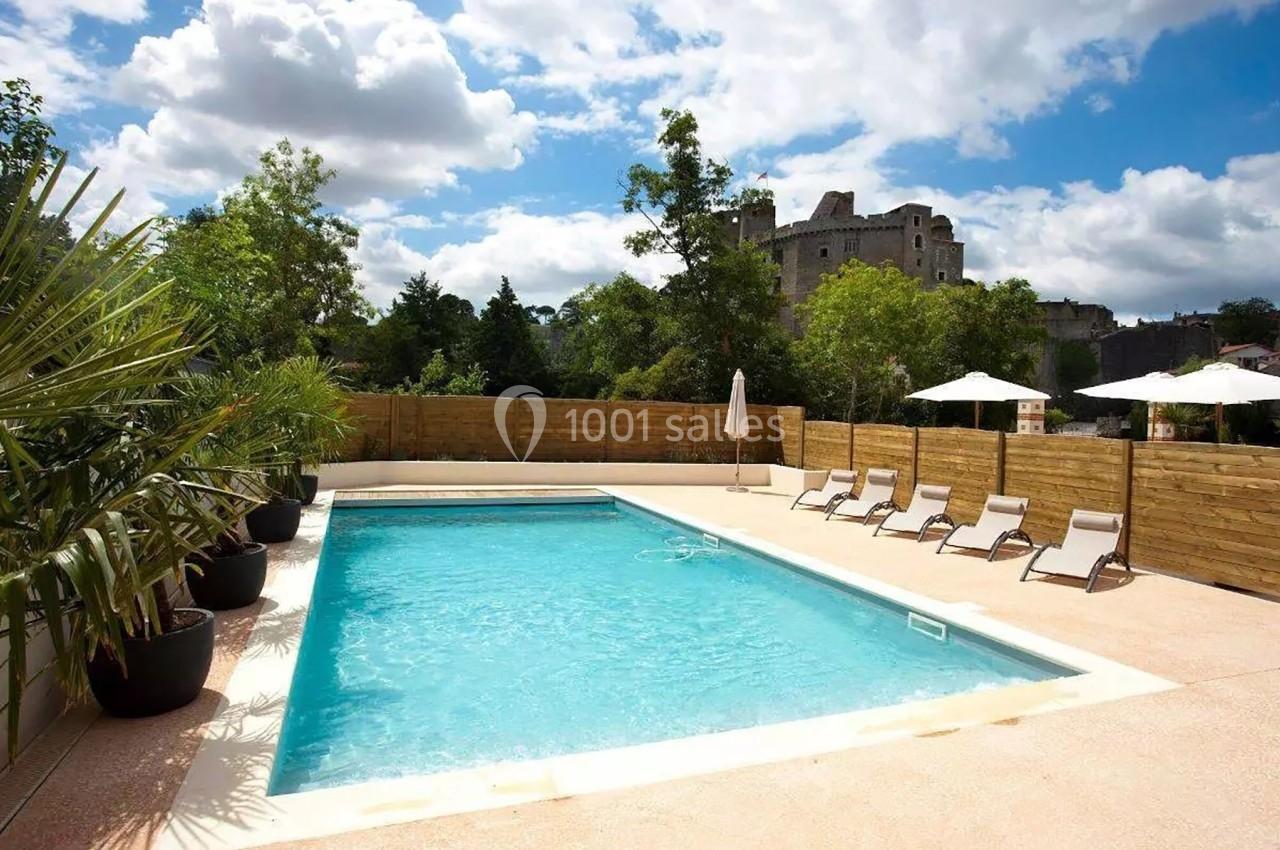 Piscine extérieure entourée de transats, parasols, palmiers et clôture en bois, avec un château en arrière-plan.