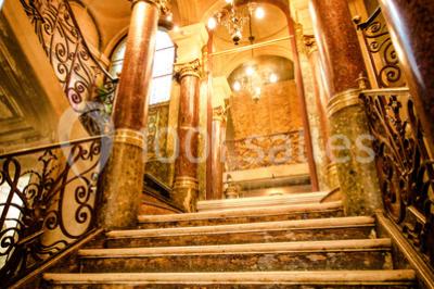 Escalier en marbre avec rampes en fer forgé, colonnes décoratives et lustres suspendus dans un intérieur ancien.