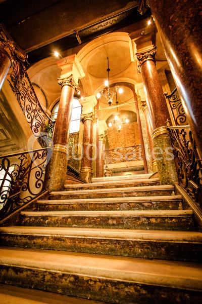 Escalier en marbre avec rampes en fer forgé, colonnes décoratives et lustres suspendus dans un intérieur ancien.