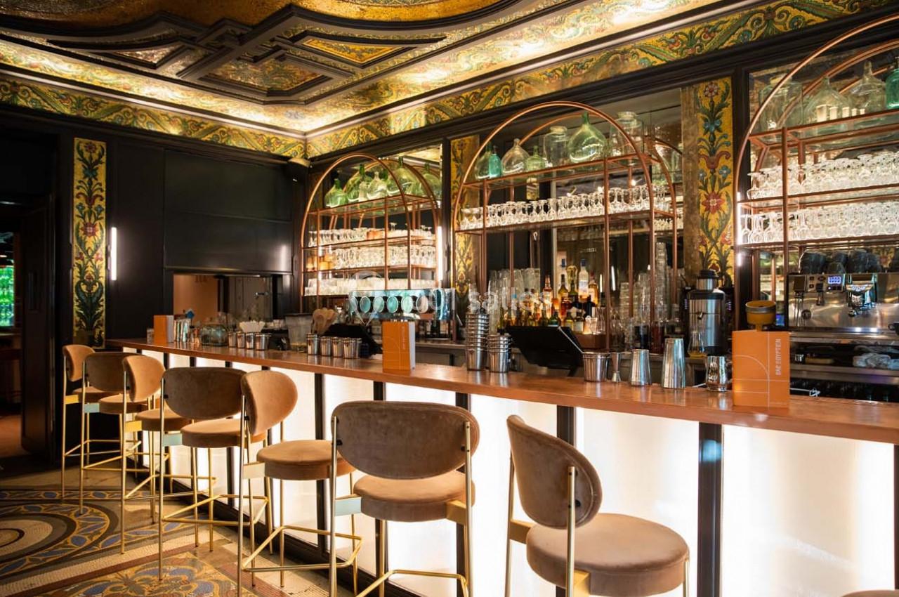 Bar élégant avec comptoir lumineux, tabourets rembourrés, étagères garnies de verres et décorations murales ornées.