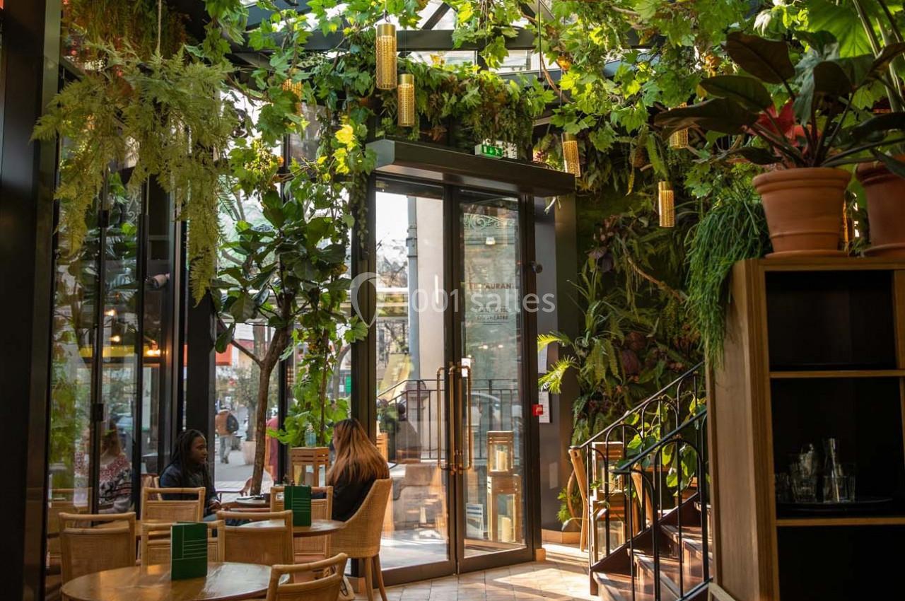 Intérieur d'un café lumineux avec des plantes suspendues, des tables en bois et des chaises en rotin.