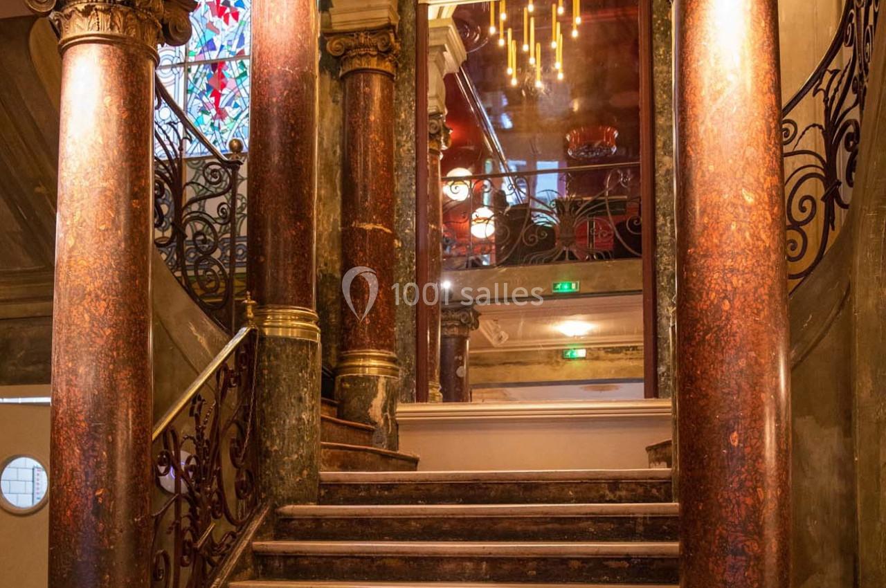 Escalier en marbre avec des colonnes décoratives et un vitrail coloré en arrière-plan, éclairé par des lustres.