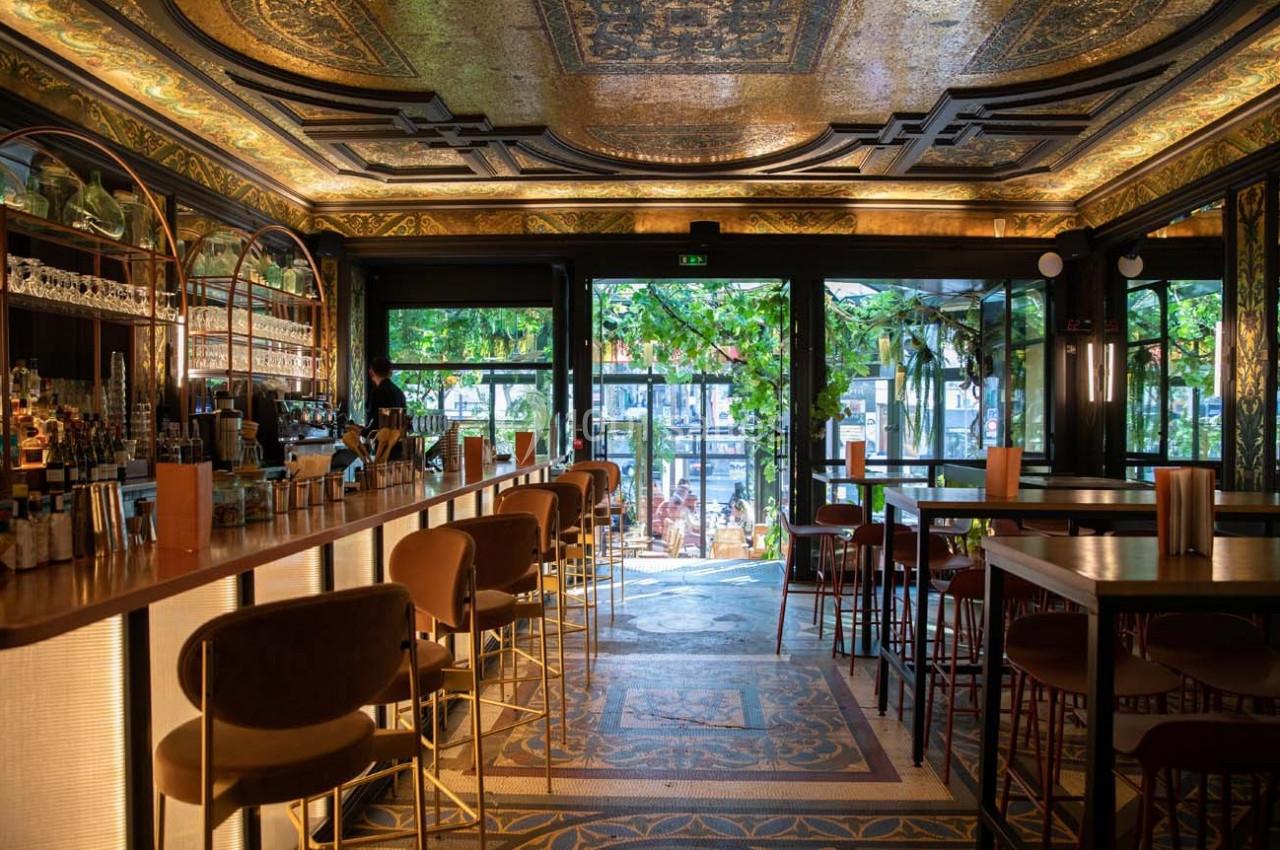 Intérieur d'un bar élégant avec un comptoir éclairé, des tables hautes et une vue sur une terrasse verdoyante.