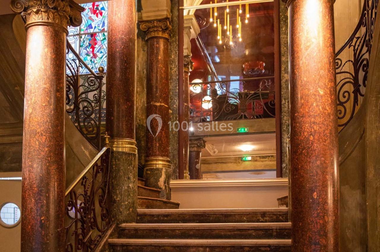 Escalier en marbre entouré de colonnes, avec une rambarde en fer forgé et un vitrail coloré en arrière-plan.