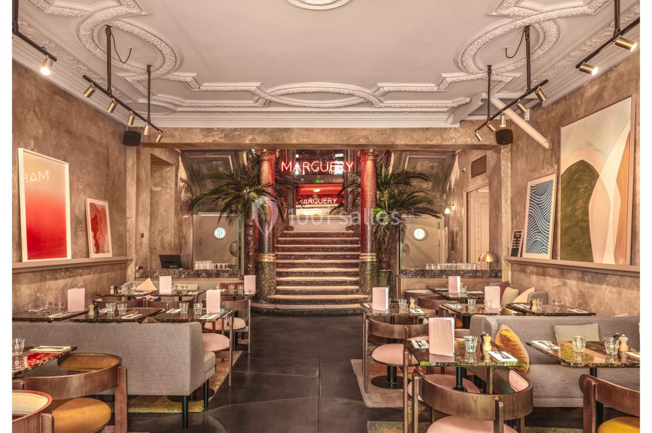 Salle de restaurant élégante avec tables dressées, décoration moderne et escalier central entouré de palmiers.